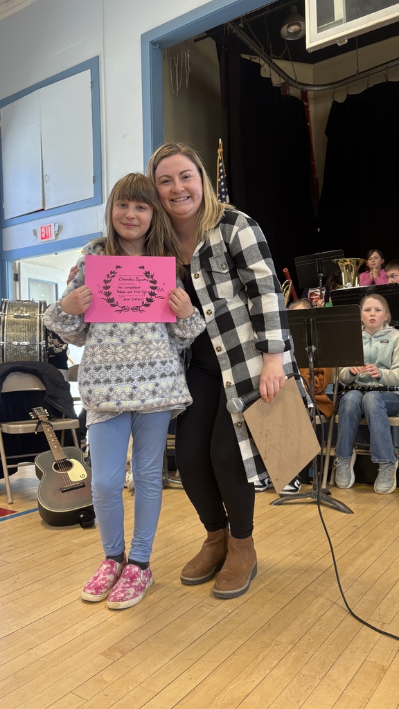 A student stands with an adult holding a bright pink certificate on a stage, with band instruments and other students visible in the background.