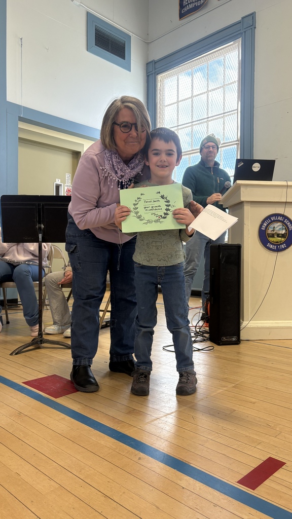 A student smiles while holding a green certificate next to an adult in a school gymnasium, with a podium and another adult speaking in the background.