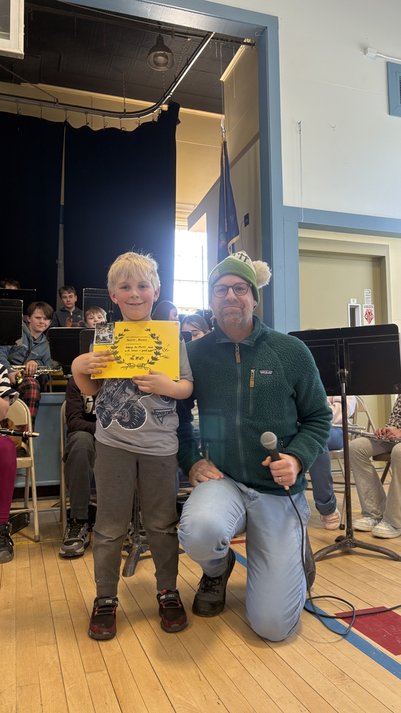 A young student stands on a school stage holding a yellow certificate next to an adult holding a microphone, with a seated student band visible in the background.