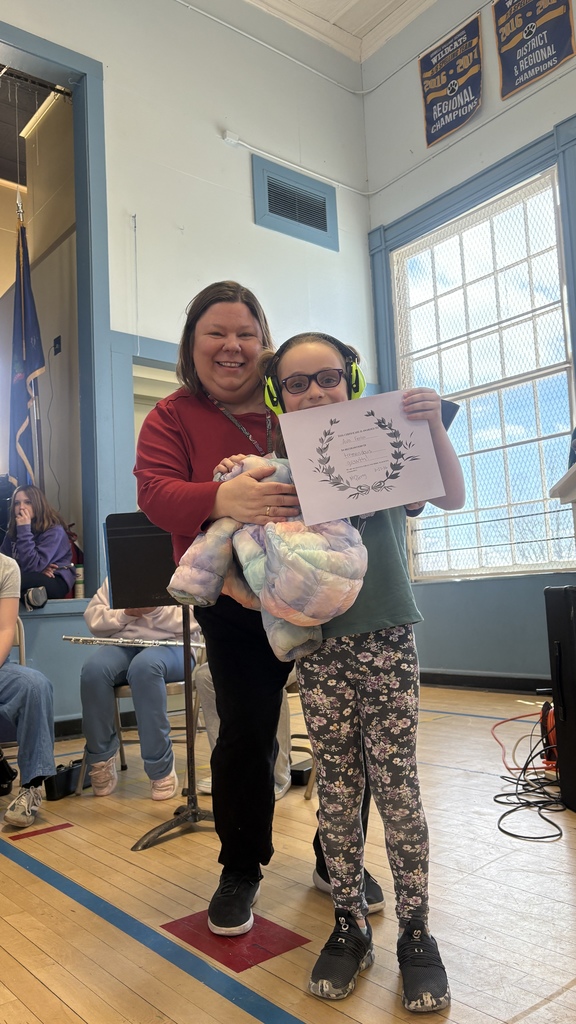 A student wearing headphones stands beside an adult and holds a certificate on a school stage, with musical instruments and students seated behind them.