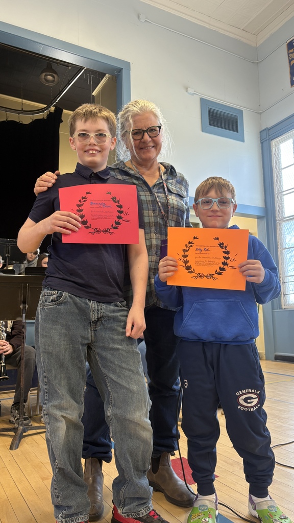 Two students stand with an adult, each holding colorful certificates (red and orange), posing on a school stage with a band setup behind them.