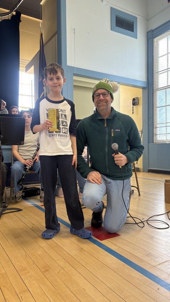 A student stands on a school stage holding a small gold-colored award next to an adult holding a microphone, with seated band students and instruments in the background.
