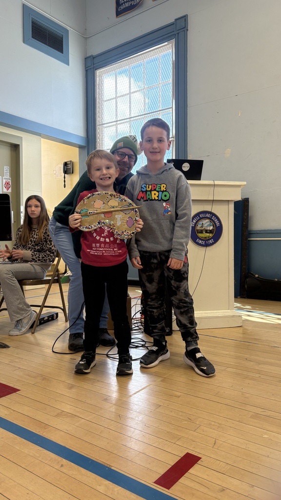 Two young students stand together smiling, one holding a decorated artist palette, while an adult stands behind them near a podium on a school stage.