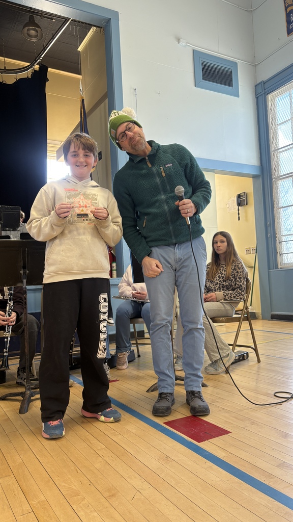 A student stands beside an adult holding a microphone and displays a small certificate or card, with other students and instruments visible behind them.