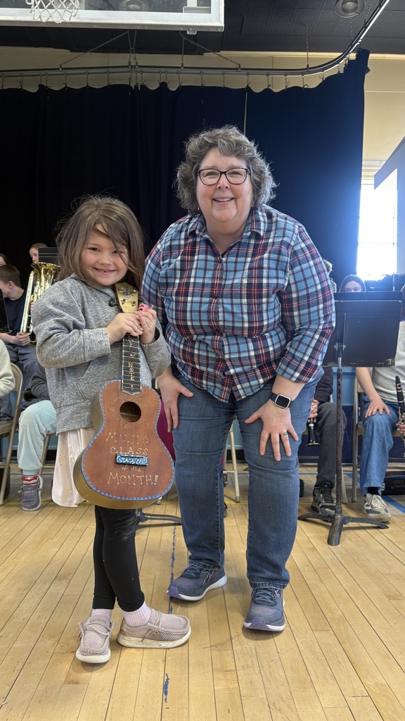 A student stands next to an adult and holds a large guitar-shaped sign that reads “Music Class of the Month,” with a school band seated in the background.