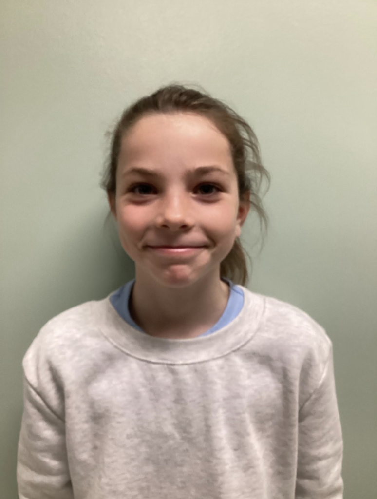 A student smiles at the camera while standing against a plain wall, wearing a light gray sweatshirt.