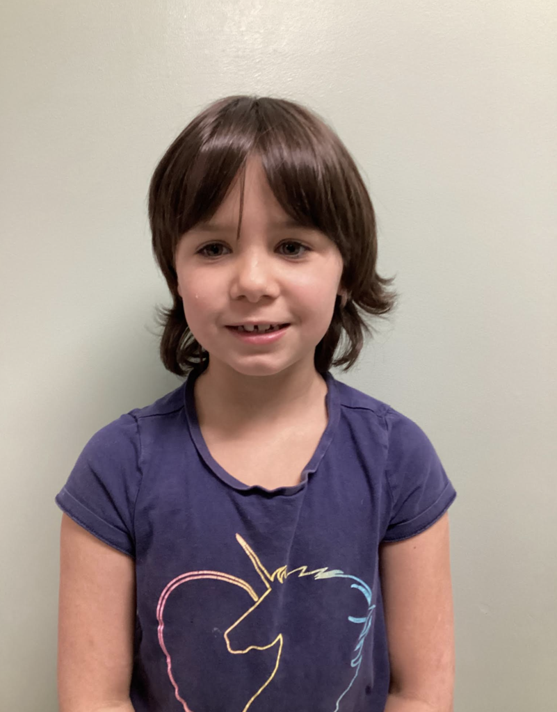A student stands against a wall wearing a purple shirt with a unicorn graphic, looking at the camera with a slight smile.