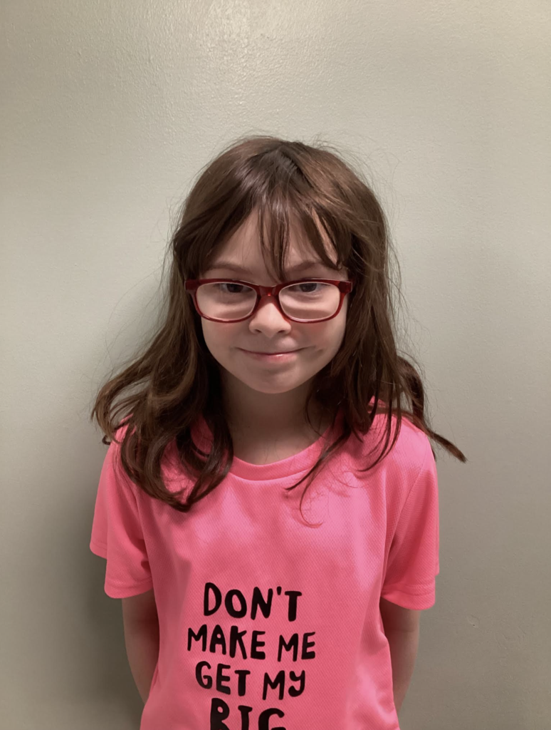 A student with red glasses and long hair stands against a wall wearing a bright pink shirt that reads, “Don’t make me get my big…”