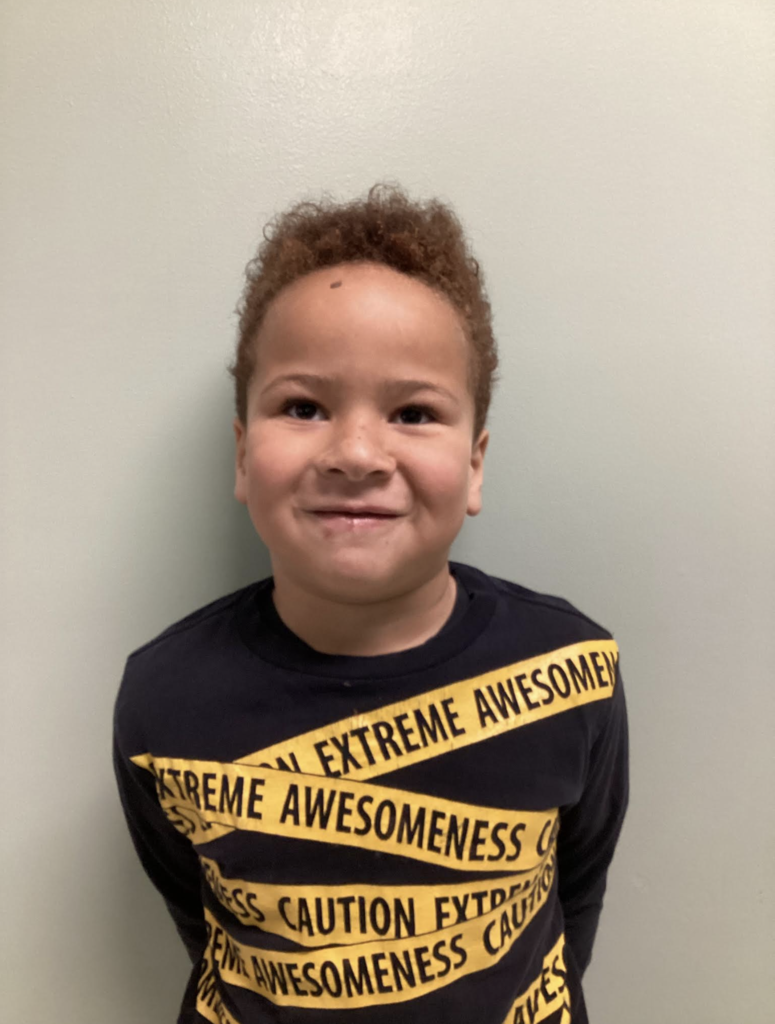 A smiling student stands against a light-colored wall wearing a black shirt with yellow caution-tape-style text that reads “Extreme Awesomeness.”