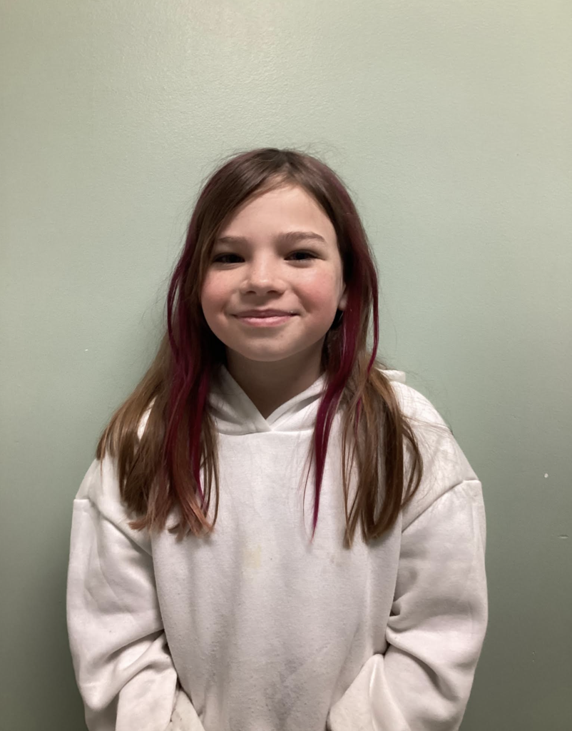 A smiling student with long hair featuring pink streaks stands against a wall wearing a white hoodie.