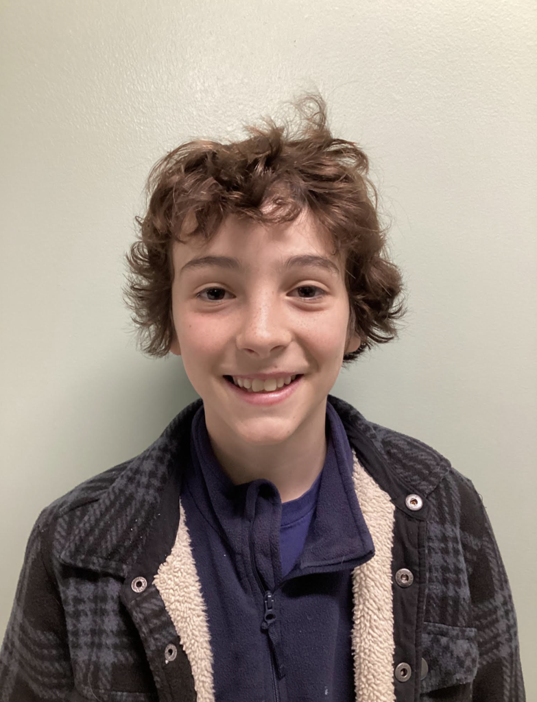 A smiling student with short, curly hair stands against a plain wall wearing a dark jacket layered over a zip-up top.