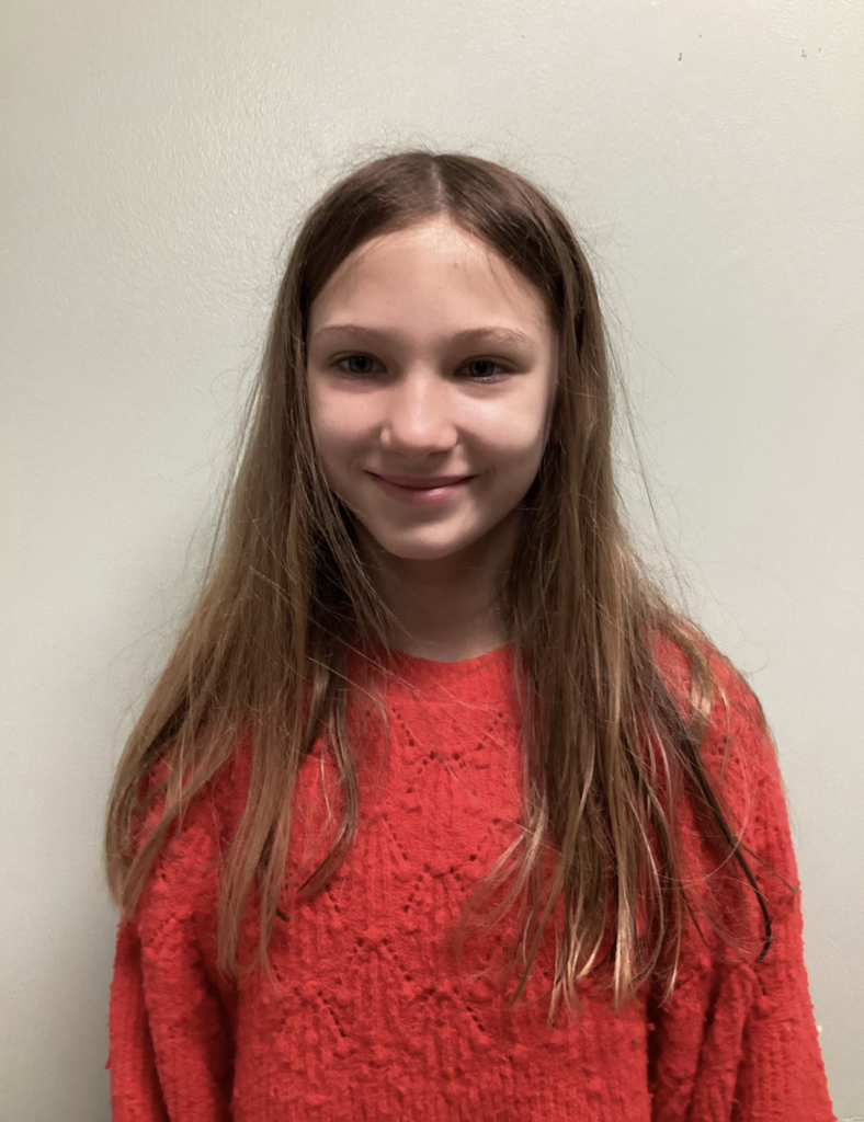 A student with long hair stands against a plain wall wearing a textured red sweater and smiling at the camera.