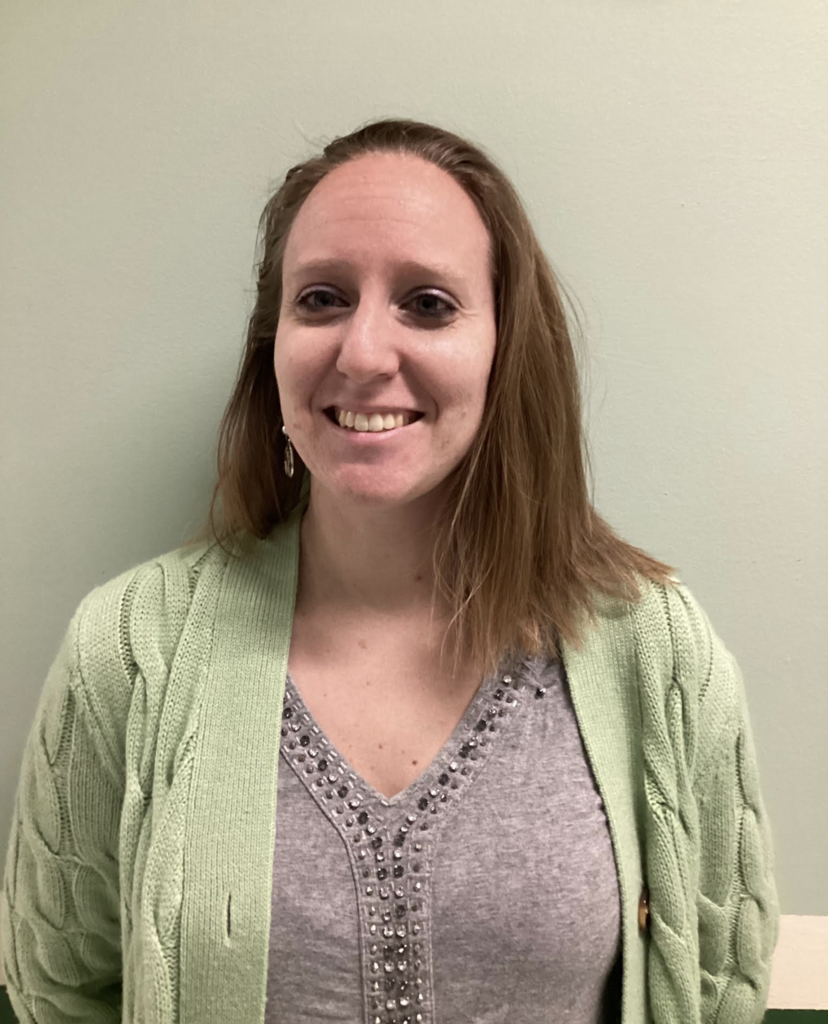 A smiling adult stands against a light-colored wall, wearing a gray top with decorative detailing and a light green cardigan.