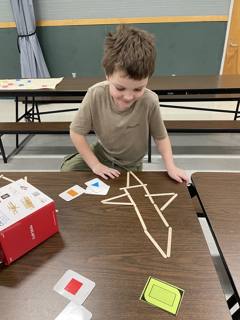   A young boy in a tan t-shirt smiles as he looks down at a table where he has used several wooden craft sticks to build a large, complex geometric shape resembling a rocket. Various colored shape flashcards are scattered around his project. 