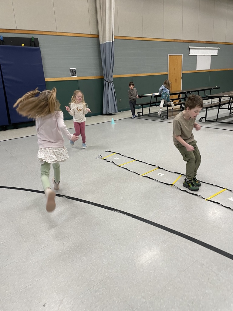 A high-energy shot of several children playing in the gymnasium. One boy is jumping through a yellow and black agility ladder laid out on the floor, while two girls run and dance nearby.