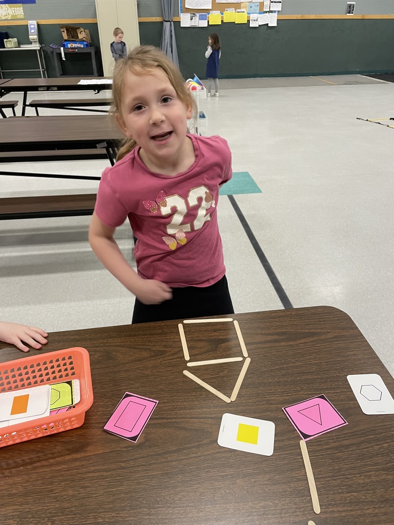  A young girl in a pink shirt stands proudly behind a table where she has built a house shape using wooden craft sticks. Several pink and white shape cards, including a triangle and a rectangle, are laid out on the table.  