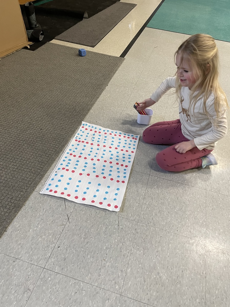   A young girl kneels on the gym floor next to a large sheet of paper covered in a grid of red and blue dots. She is holding several colored pencils, and a large blue die sits on the floor nearby.  