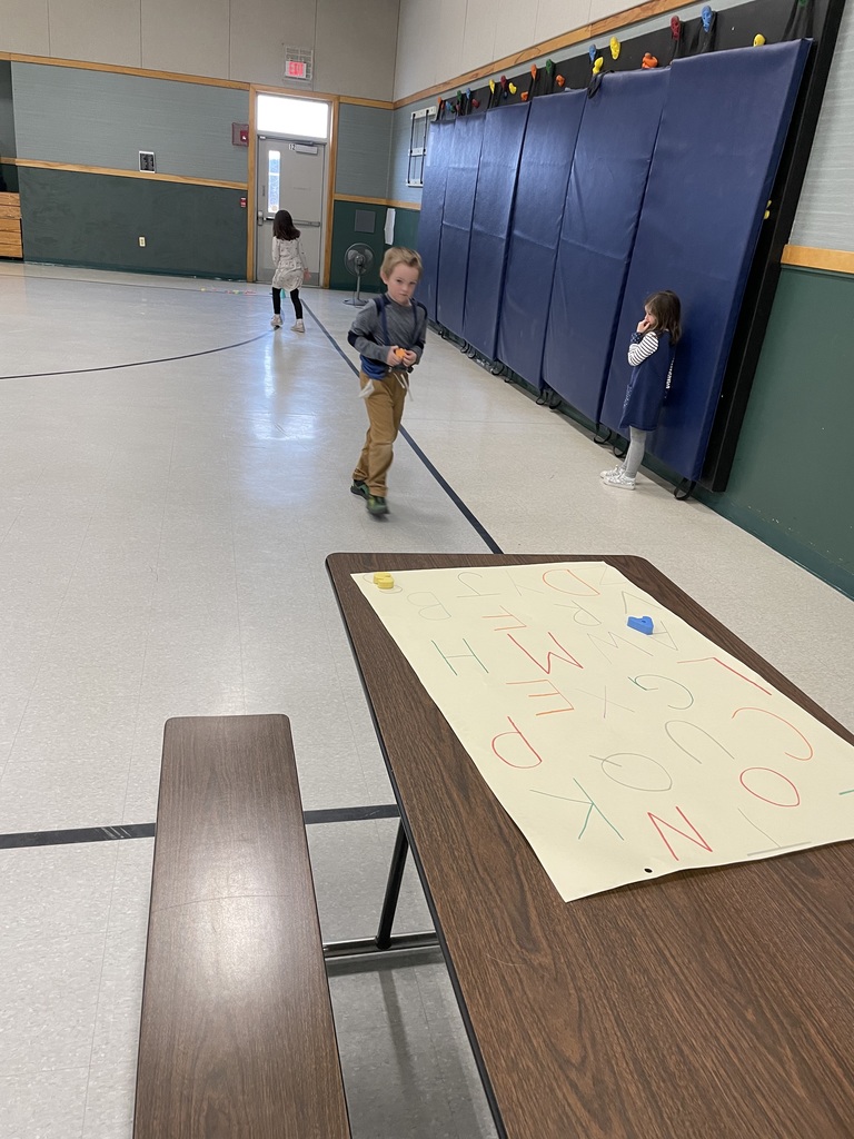 A wide shot of a school gymnasium featuring a large paper alphabet poster on a brown folding table in the foreground. In the background, three young children are moving around the gym, which has green and grey walls and blue safety mats along one side.  
