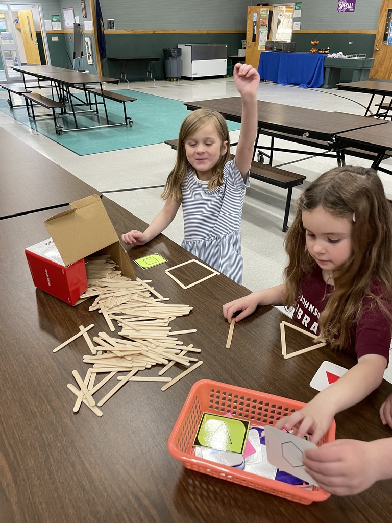 Two young girls work at a table with a large pile of wooden craft sticks. One girl has her hand raised excitedly while the other focuses on building a triangle. They are using flashcards with shapes like squares and hexagons as guides. 