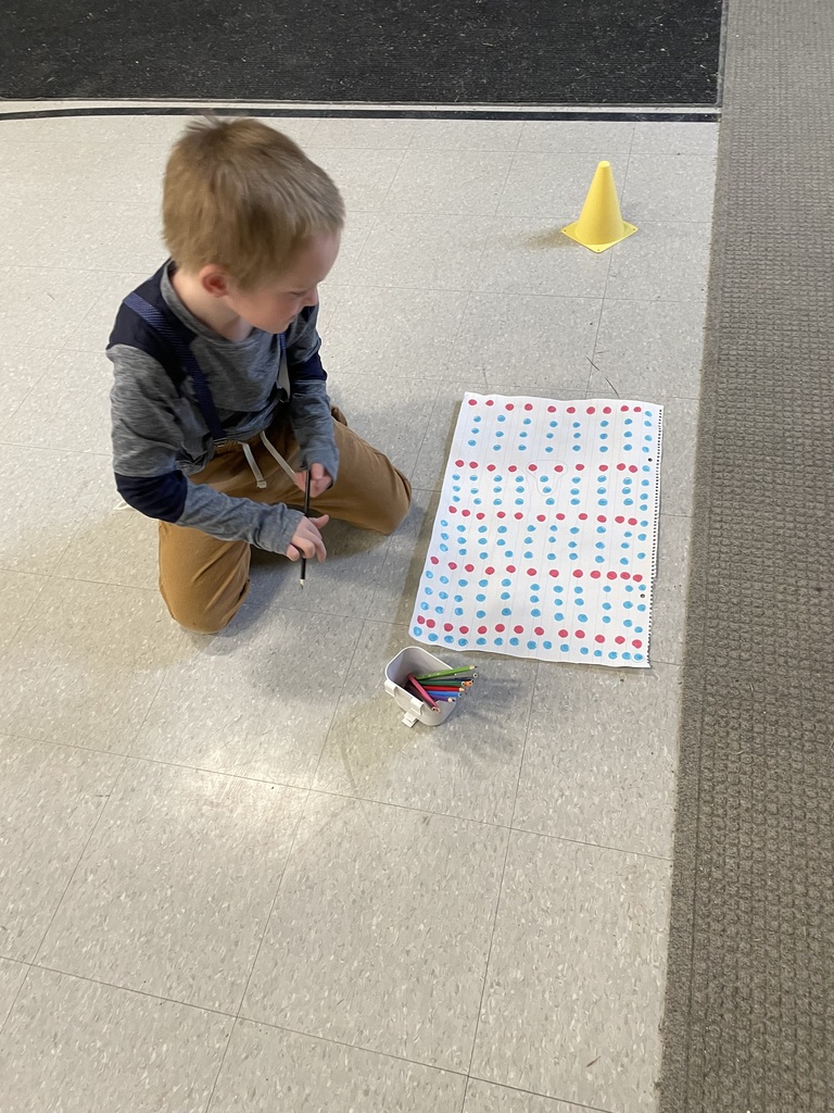  A young boy in a tan t-shirt smiles as he looks down at a table where he has used several wooden craft sticks to build a large, complex geometric shape resembling a rocket. Various colored shape flashcards are scattered around his project.  