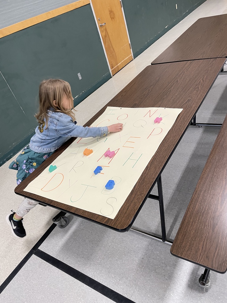   A young girl in a denim jacket leans over a table to match colorful 3D foam letters to hand-drawn letters on a large white poster. Several foam letters like 'M', 'F', and 'K' are already placed on their corresponding spots.  📐 Shape & Geometry Stations