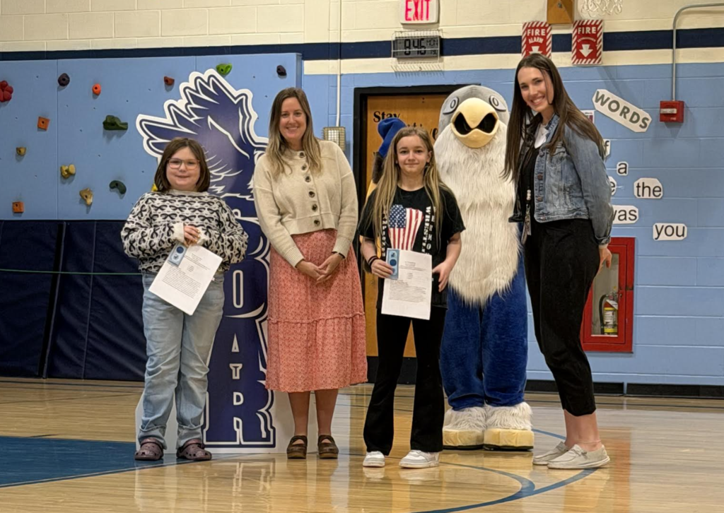 Two adults and two students stand in a gym holding certificates near a hawk “Soar” display, with the school mascot behind them.