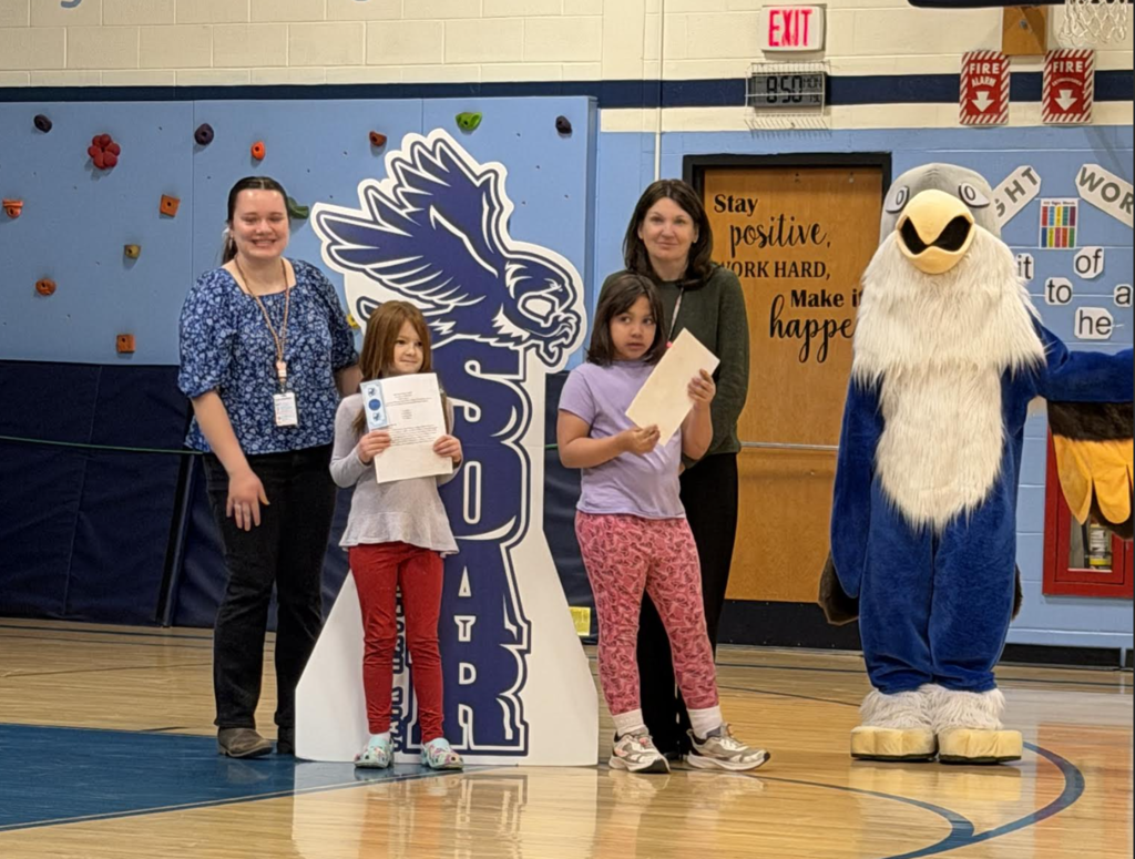 Two adults and two students pose in a gym holding certificates next to a “Soar” hawk sign, with a hawk mascot standing to the side.