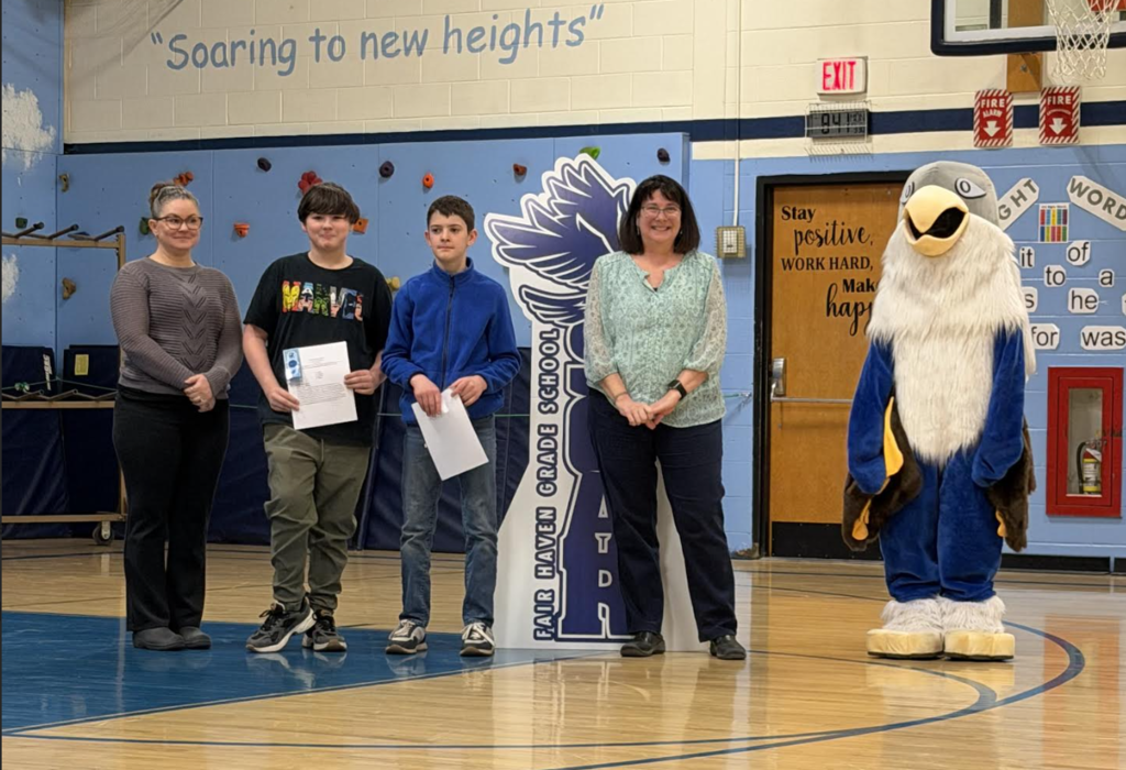 Two adults and two students stand in a school gym holding certificates beside a “Soar” hawk sign and eagle mascot.
