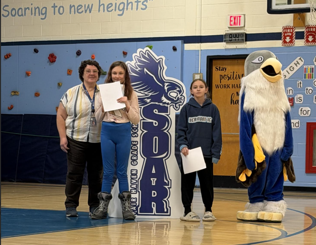 An adult and two students stand in a school gym holding certificates beside a large “Soar” hawk sign, with a school mascot in an eagle costume nearby.