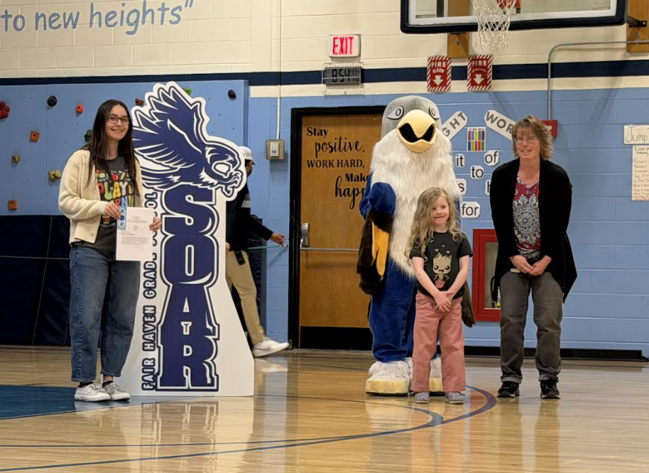 An adult and a young student stand in a school gym holding a certificate beside a “Soar” hawk display, with the school mascot nearby.