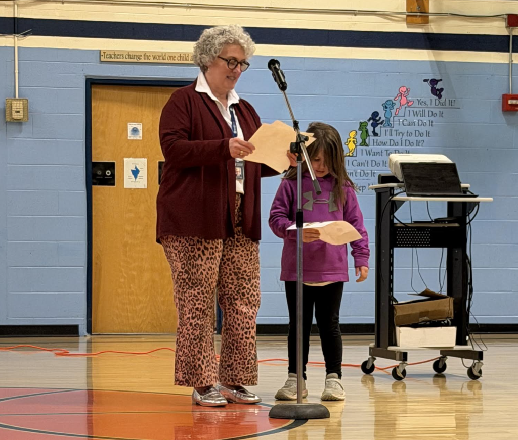 An adult and a young student stand at a microphone in a gym, reading from papers during a school assembly.