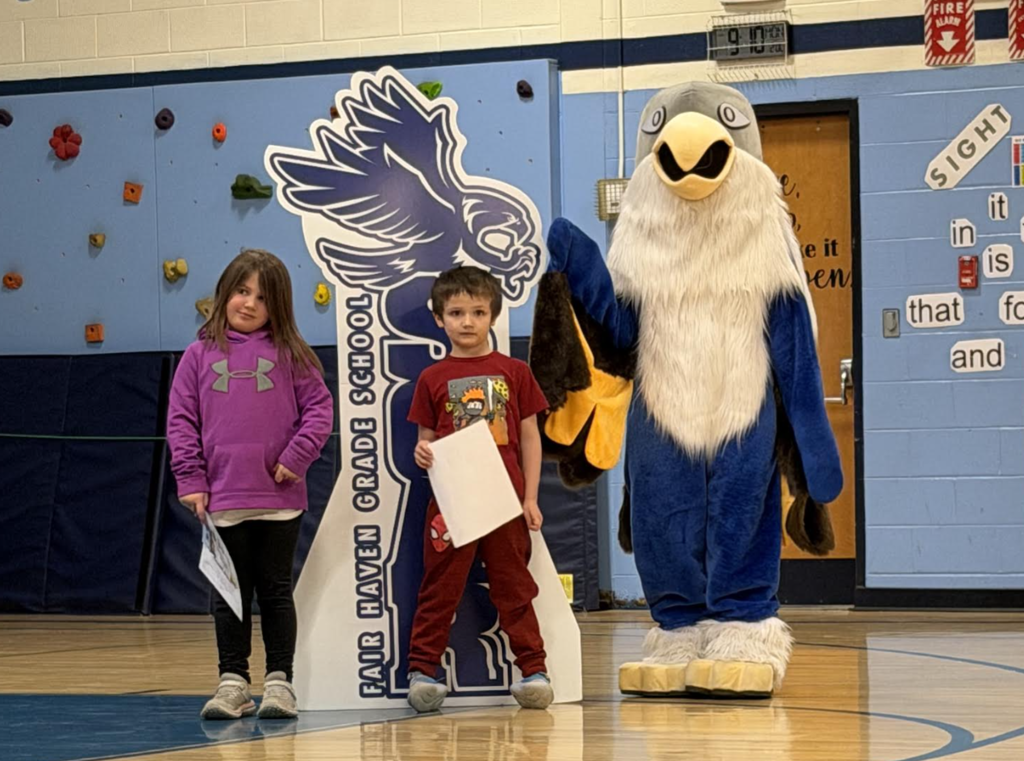 Two young students stand in a gym holding certificates next to a “Soar” hawk sign, with the eagle mascot behind them.