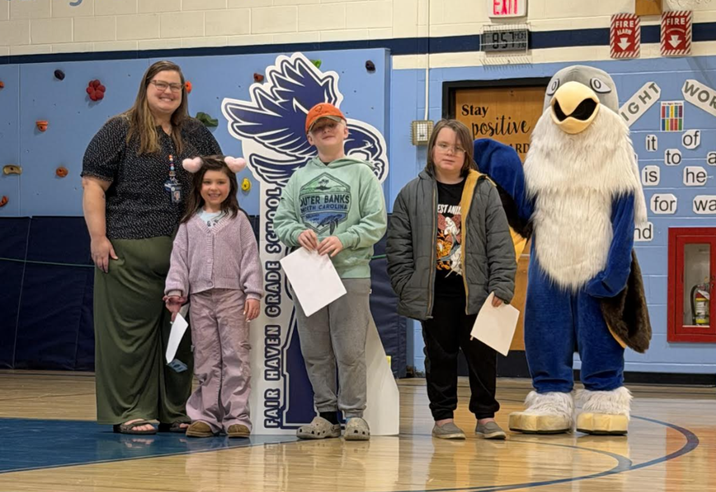 An adult stands with three students in a gym, two holding certificates, next to a “Soar” hawk sign and eagle mascot.