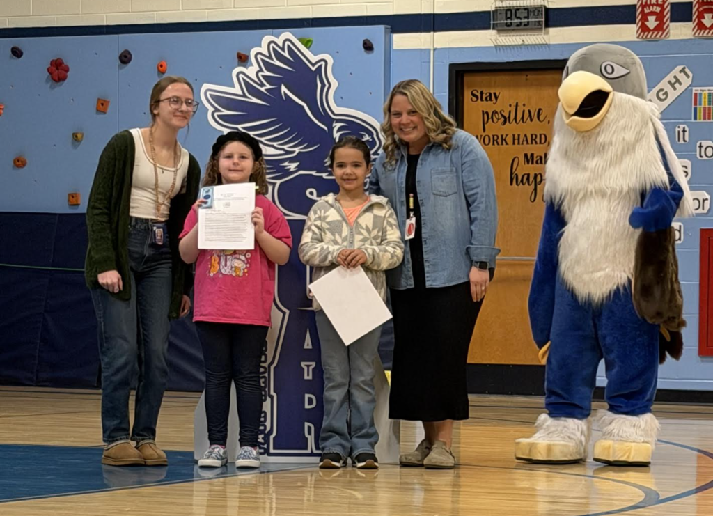 Two adults and two students pose in a gym holding certificates beside a hawk “Soar” display, with the school mascot standing to the side.