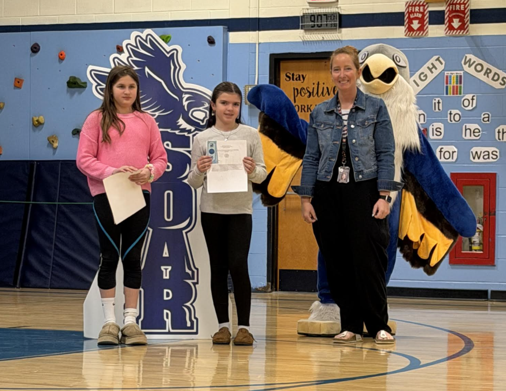 Two students hold certificates while standing with an adult in a school gym beside a “Soar” hawk display and eagle mascot.