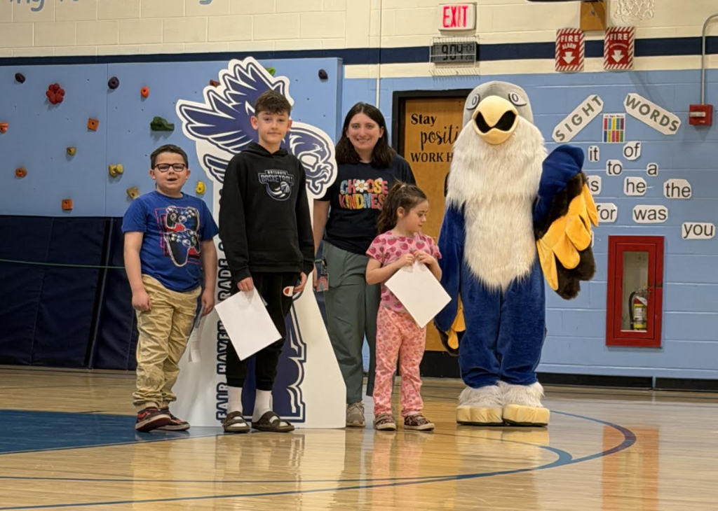 An adult and three students stand in a gym holding certificates beside a hawk “Soar” display, with the school mascot nearby.