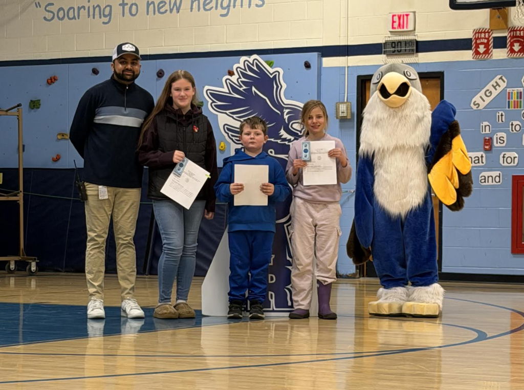 An adult and three students pose in a school gym holding certificates next to a “Soar” hawk sign and eagle mascot.