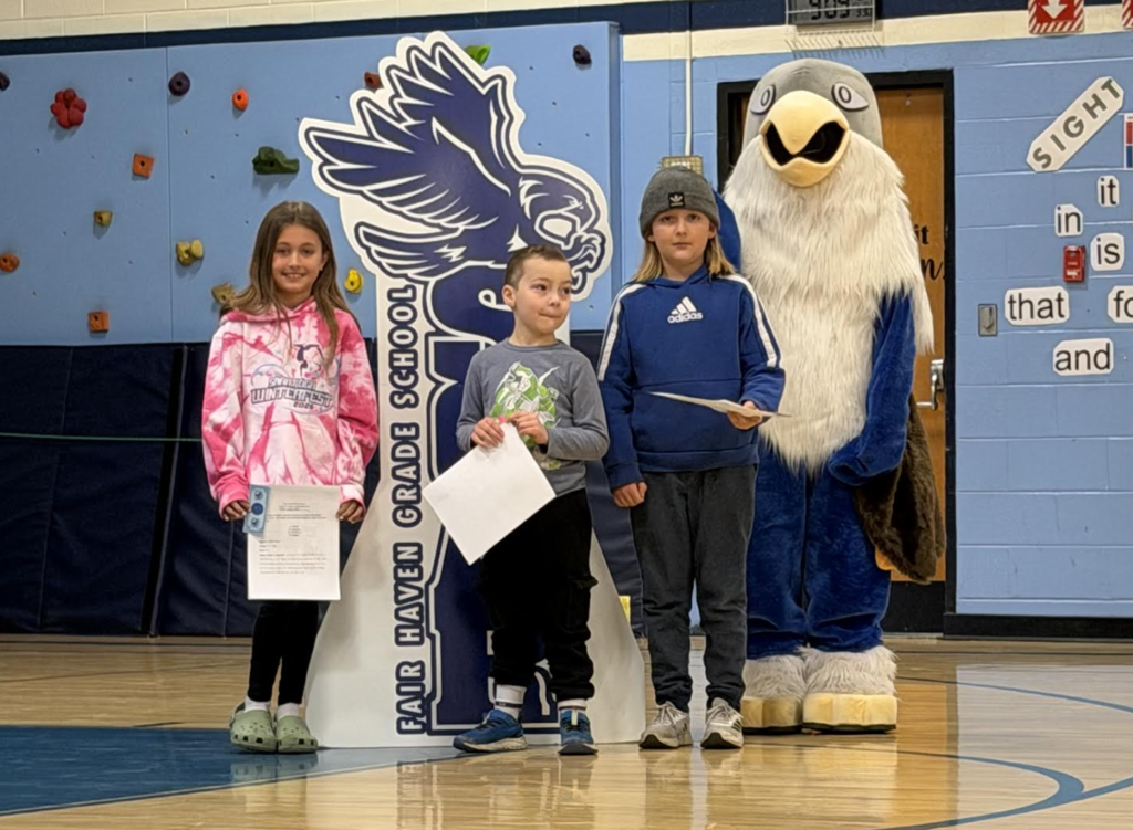 Three students pose in a gym holding certificates next to a “Soar” hawk sign, with the eagle mascot behind them.
