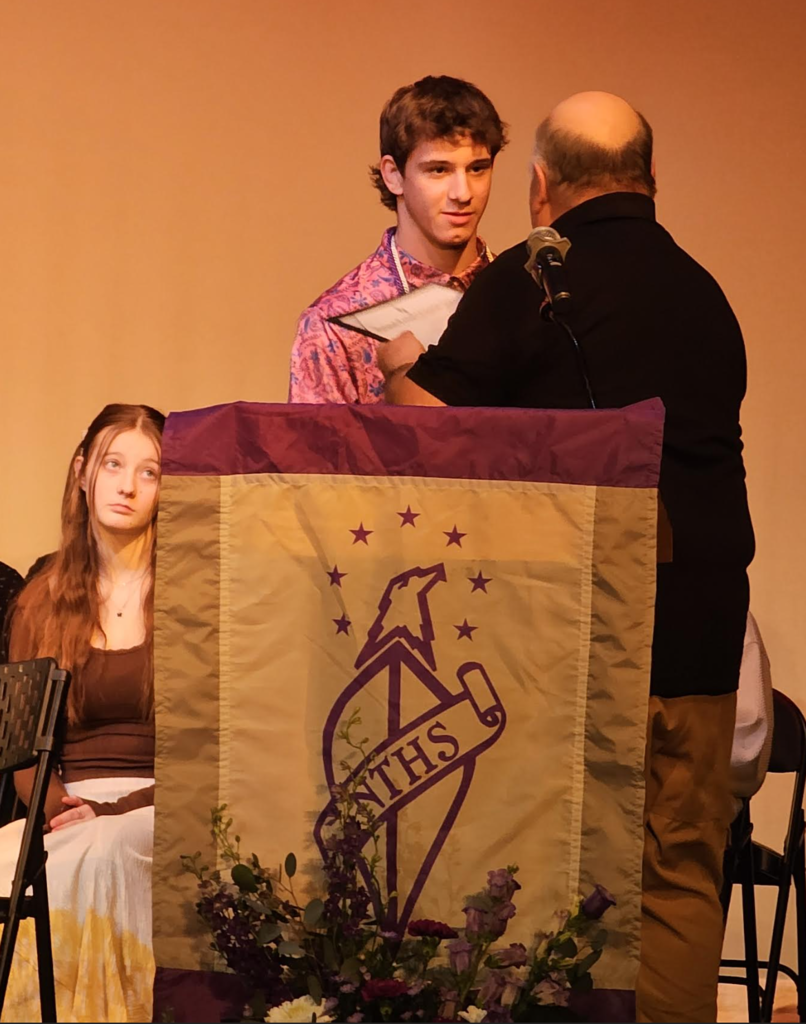 Student stands on stage receiving a certificate during a National Technical Honor Society induction ceremony, with a podium and banner in the foreground.