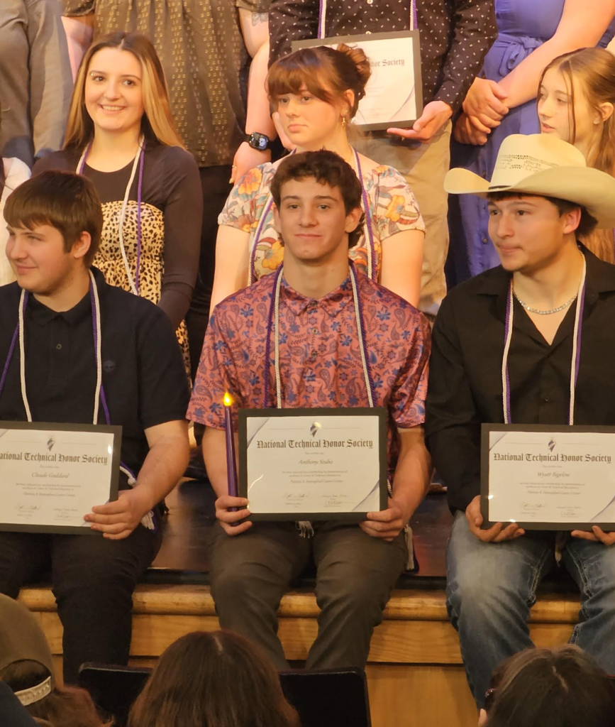 Group of students seated on stage holding National Technical Honor Society certificates, including one student in the center wearing a patterned shirt and honor cords, posing for a recognition photo.