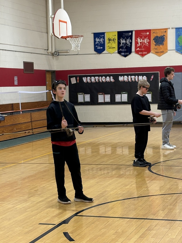 Students stand spaced apart in a gym practicing fly casting techniques with rods, focused on their movements.