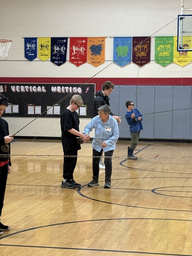 Instructor assists two students in a gym as they practice holding fly fishing rods, with others learning nearby.