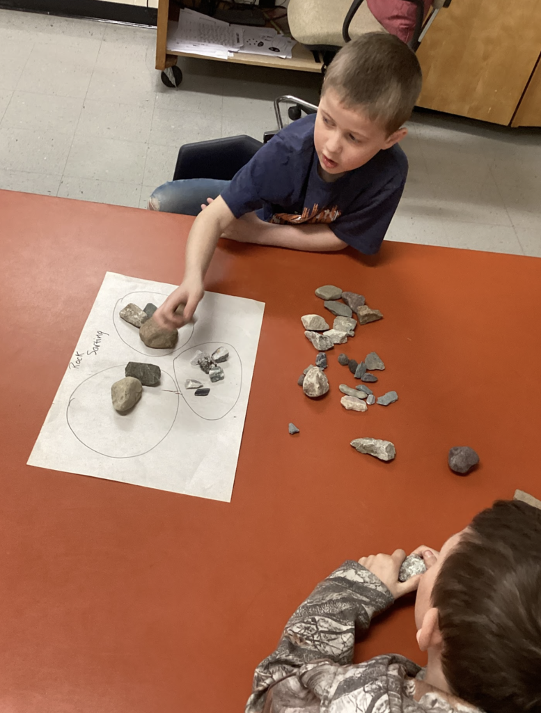 Student reaches to place a rock into a labeled group on paper while sorting rocks by size and type at a table.