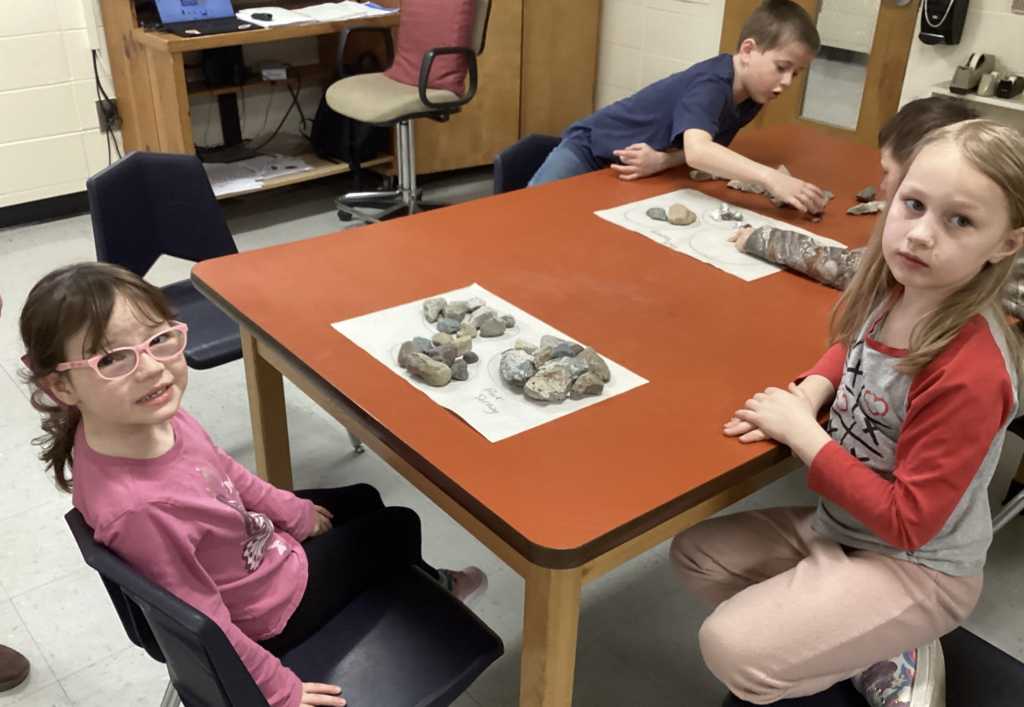 Two students sit at a table with piles of rocks sorted into groups on paper, looking toward the camera.