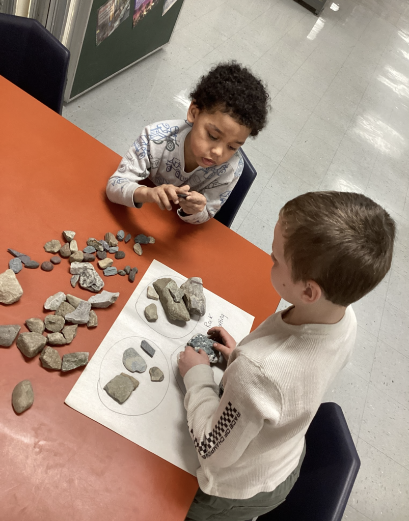 Two students sit at a table sorting a variety of rocks into groups on paper based on different characteristics.