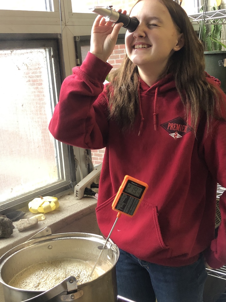 Student smiles while looking through a handheld refractometer next to a pot of boiling sap with a thermometer.