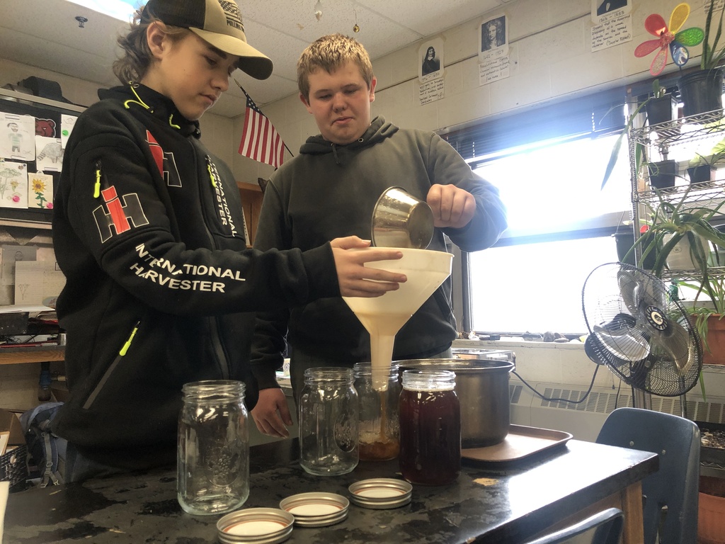 Two students pour hot maple sap through a filter funnel into glass jars in a classroom.