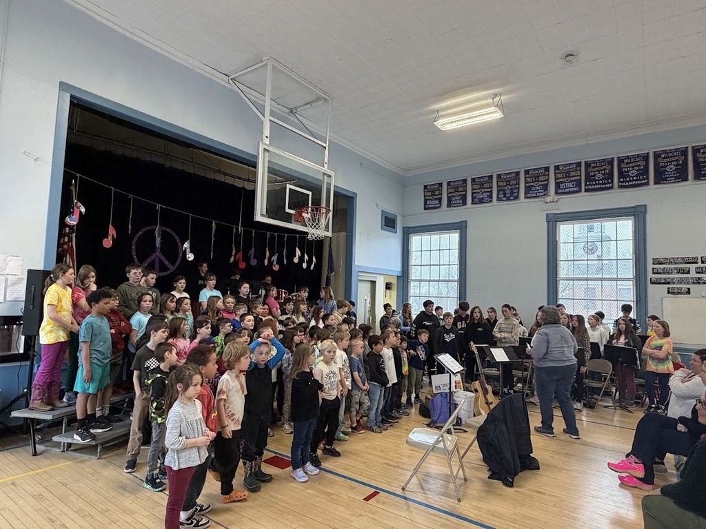 Large group of students performs together in a school gym with a conductor leading and an audience seated nearby.