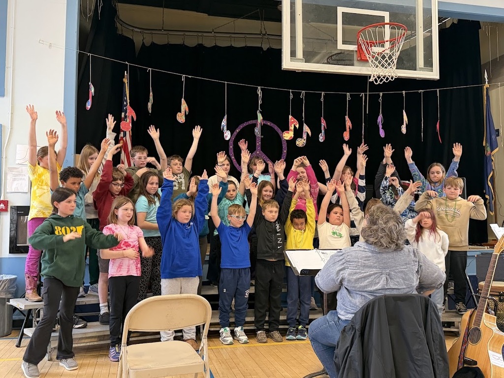 Students on risers raise their hands while singing during a school concert, led by a teacher seated in front of them.