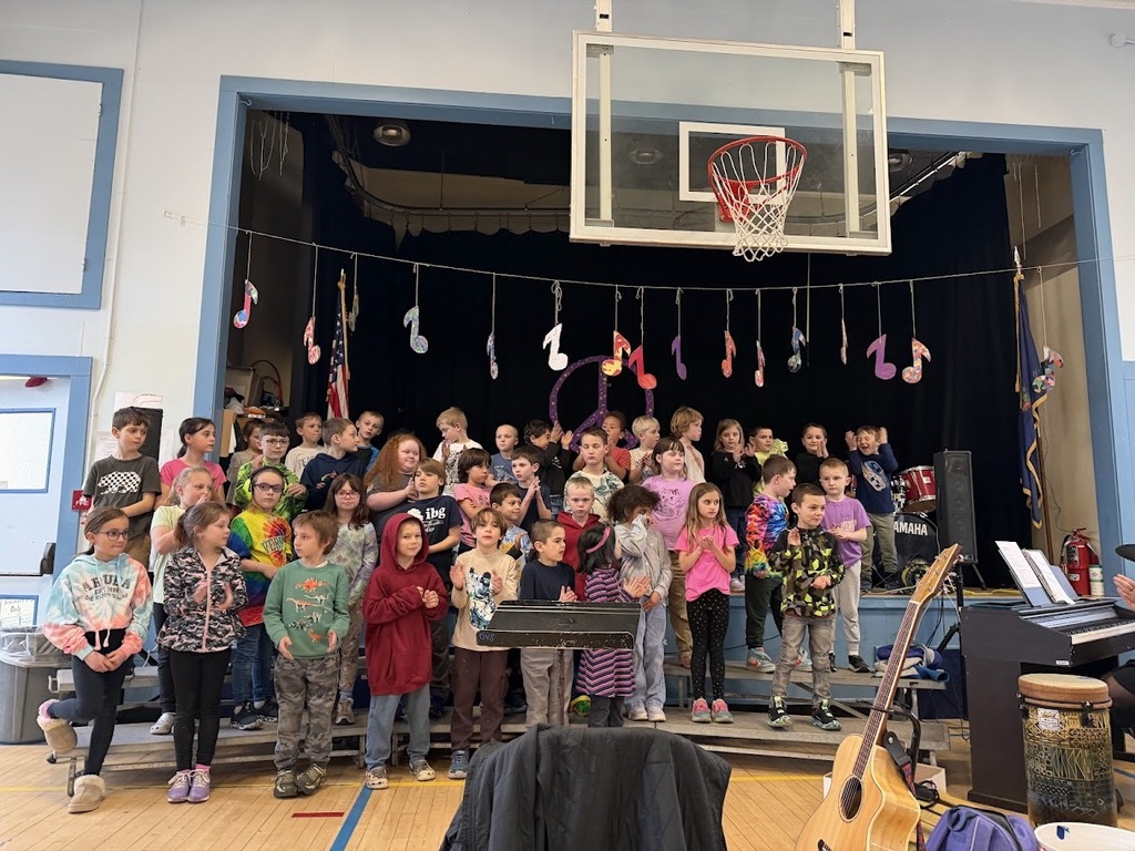 Elementary students stand on risers in a school gym performing a concert with music note decorations hanging behind them.